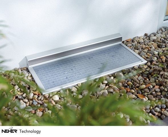 Silberne Fensterschachtabdeckung vor einer weißen Wand, eingebettet in ein Bett aus Steinen und Grün.