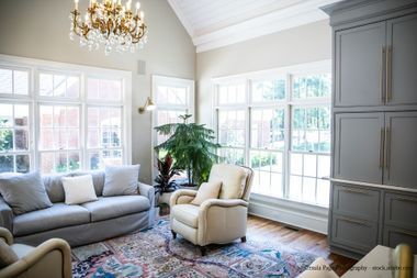 Wintergarten mit Sofa, Sessel und grauen Schränken; große Fenster, Kronleuchter und gemusterter Teppich.