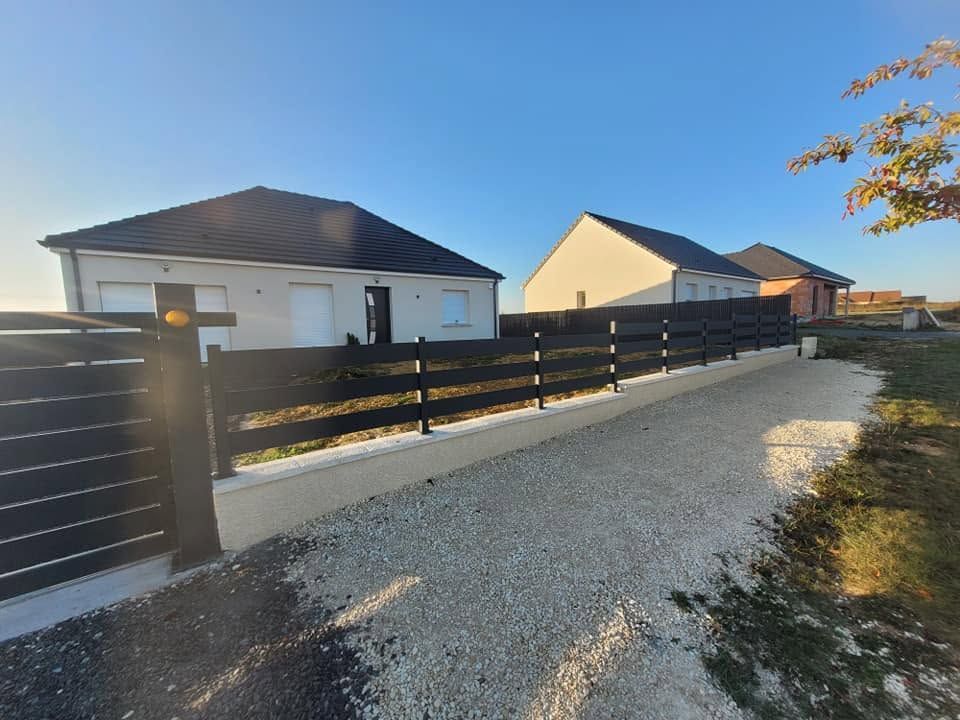 Maison moderne avec une clôture noire bordant une allée en gravier, baignée de soleil