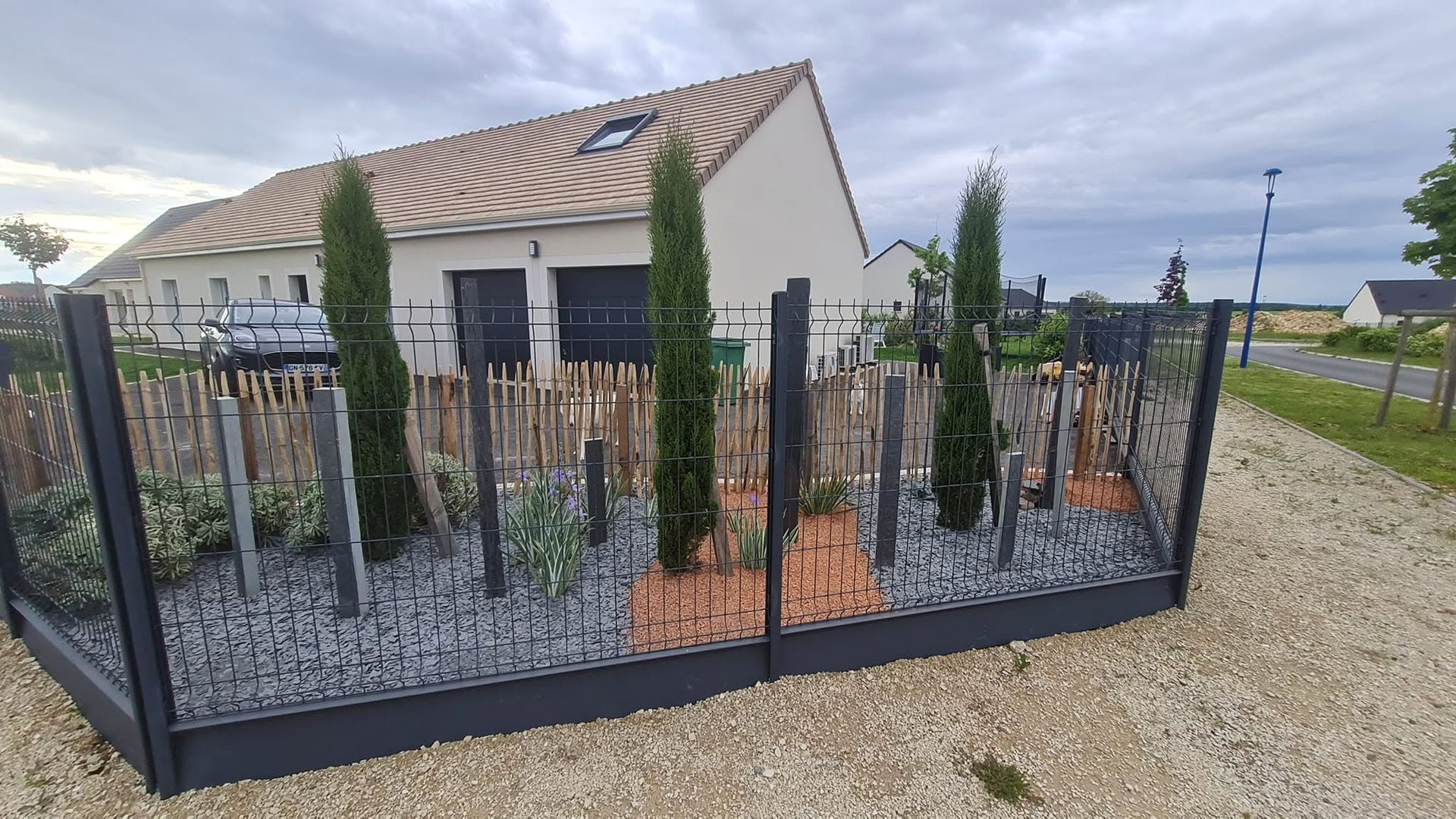 Maison moderne avec une clôture noire et un jardin paysager à l’avant, sous un ciel nuageux