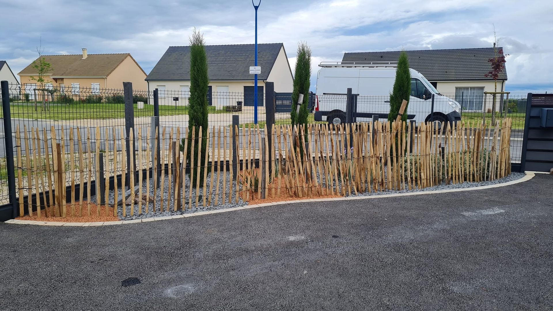 Potager clôturé avec de jeunes plants et une allée de gravier devant des maisons sous un ciel nuageux