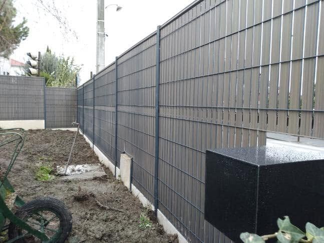 Grande clôture sombre le long d’une étroite cour latérale extérieure, au sol nu, avec un boîtier technique noir