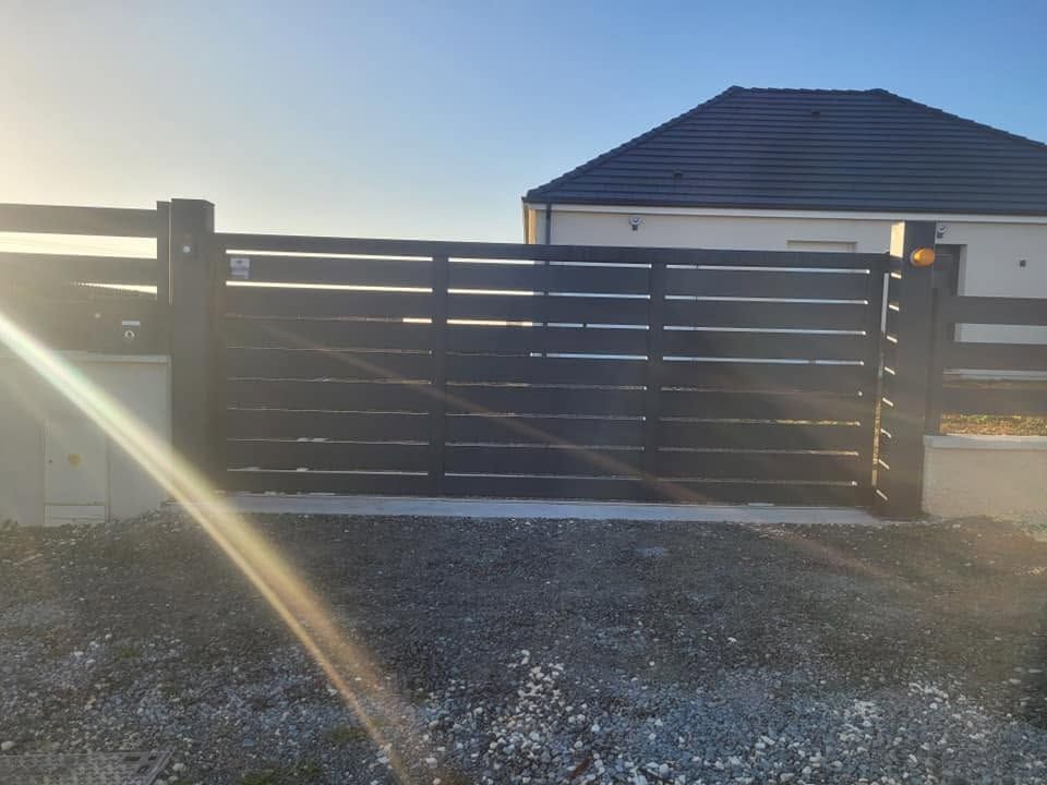 Portail coulissant noir moderne à l’entrée d’une allée en gravier, avec des reflets du soleil