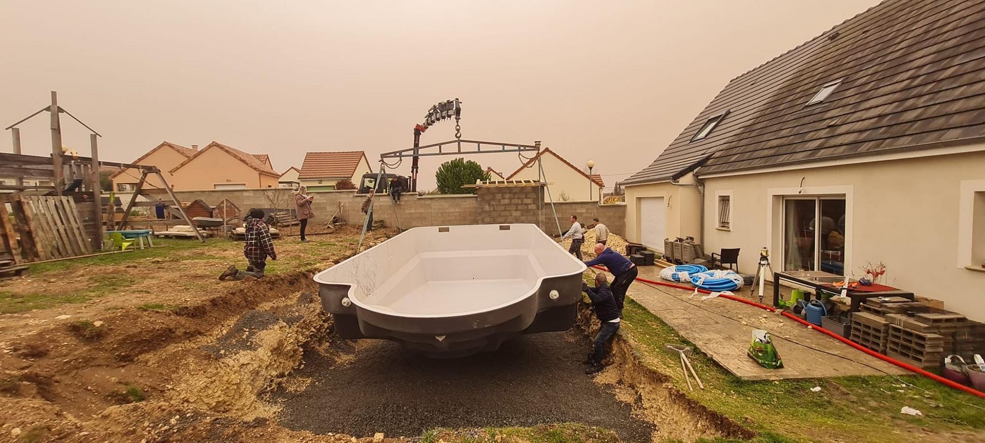 Des ouvriers déplacent la coque blanche d’un bateau dans une cour boueuse en banlieue, à côté d’une maison et de matériaux de construction