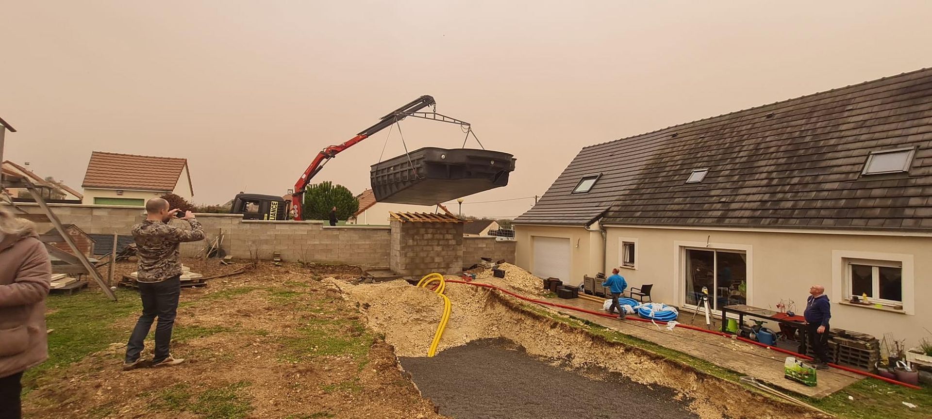 Un engin de chantier soulève de la terre à côté d’une maison en réparation, tandis que des ouvriers se trouvent à proximité et que des débris jonchent le jardin