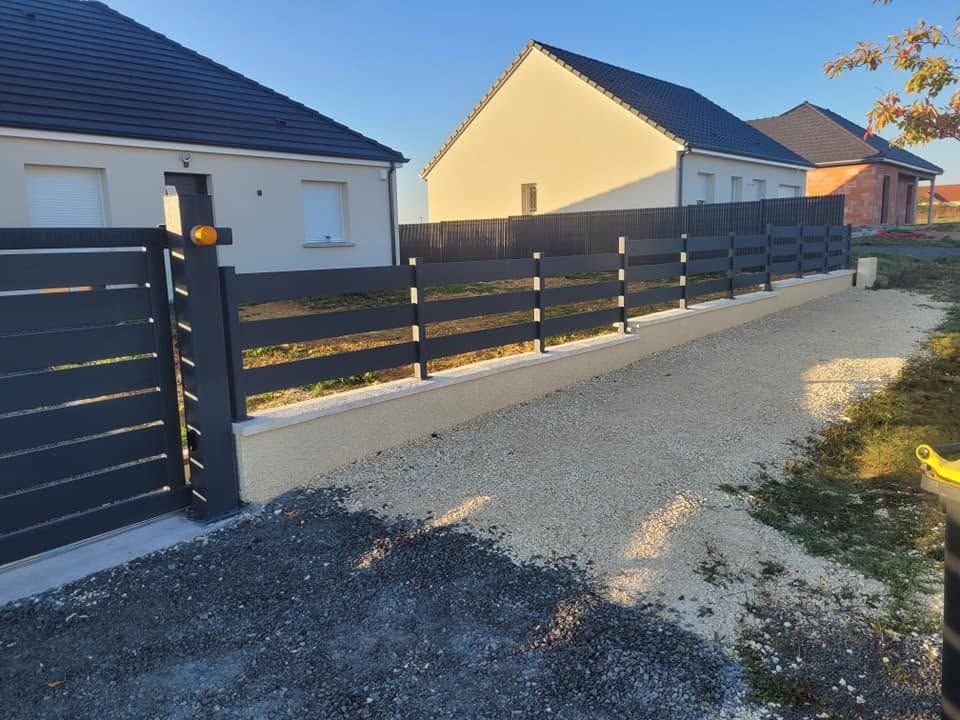 Clôture noire à lames sur un muret en béton, bordant une allée en gravier à côté de maisons modernes au coucher du soleil