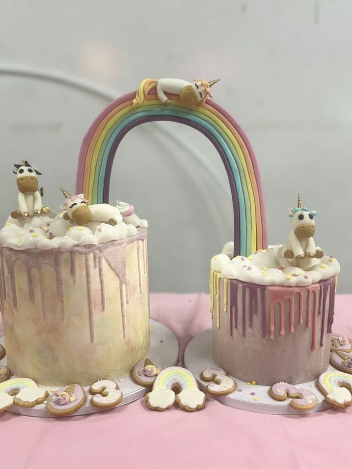 Sobre una mesa hay dos pasteles con unicornios y un arcoíris encima.