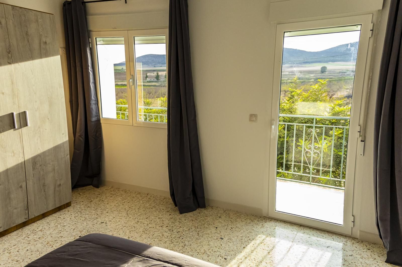 Dormitorio con ventana y balcón con vista a un paisaje rural, cortinas grises, suelo de terrazo.