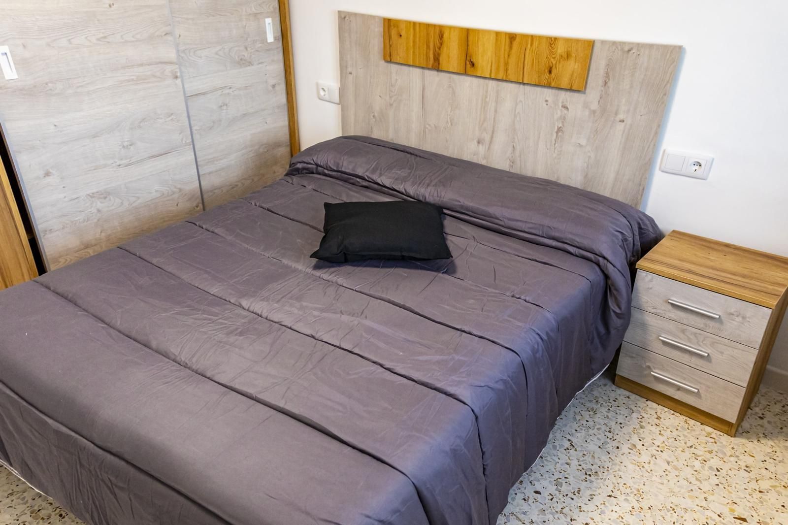 Dormitorio con cama, mesita de noche y armario. La cama tiene un edredón morado y una almohada negra.