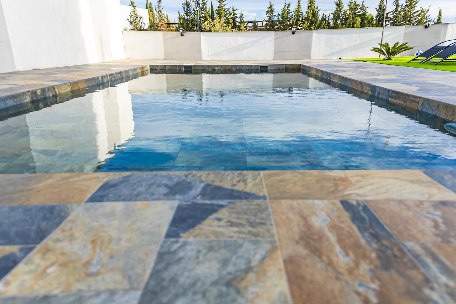Piscina rectangular con marco de baldosas de piedra, que refleja el cielo, en un entorno exterior soleado.