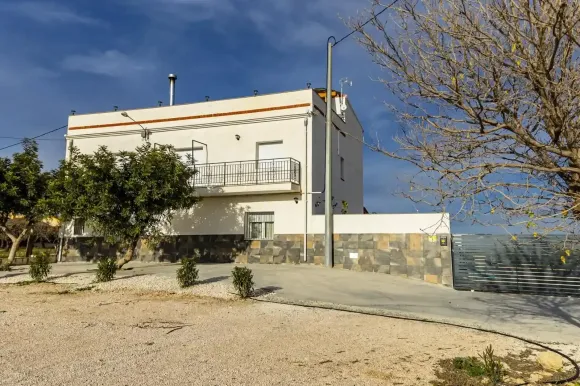 Casa blanca de dos pisos con balcón, base de piedra oscura y entrada para autos bajo un cielo azul.