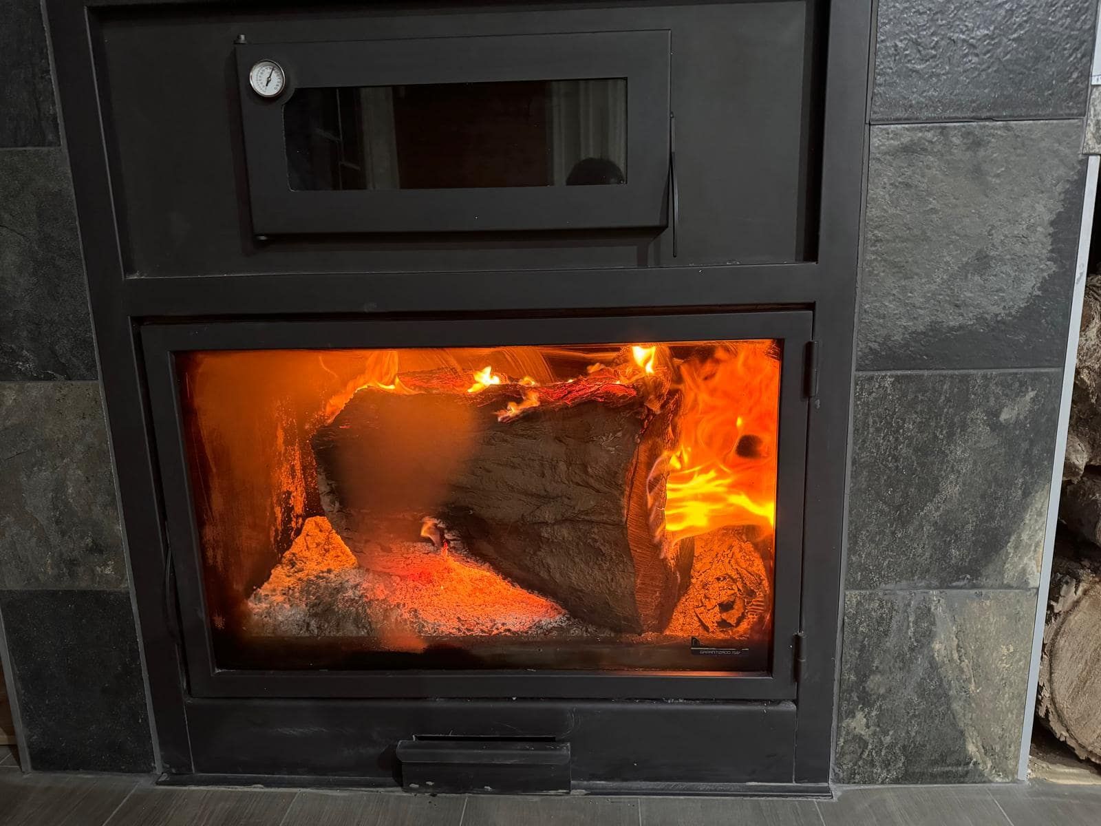 Fuego ardiendo dentro de una chimenea negra con puerta de cristal, rodeada de piedra gris.