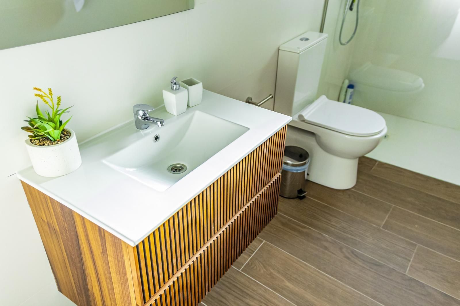 Baño con tocador de madera, lavabo y encimera blancos, inodoro y suelo imitación madera color marrón.