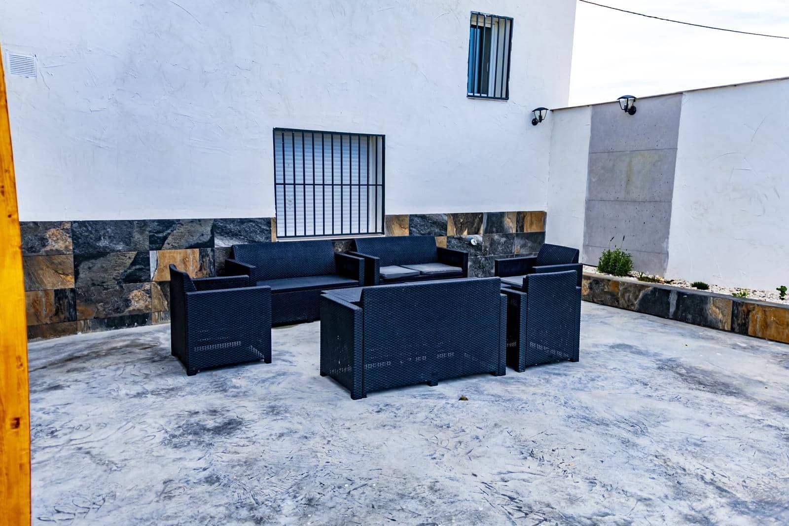 Zona de estar al aire libre con muebles de mimbre negro en un patio de hormigón. Paredes blancas y de piedra.