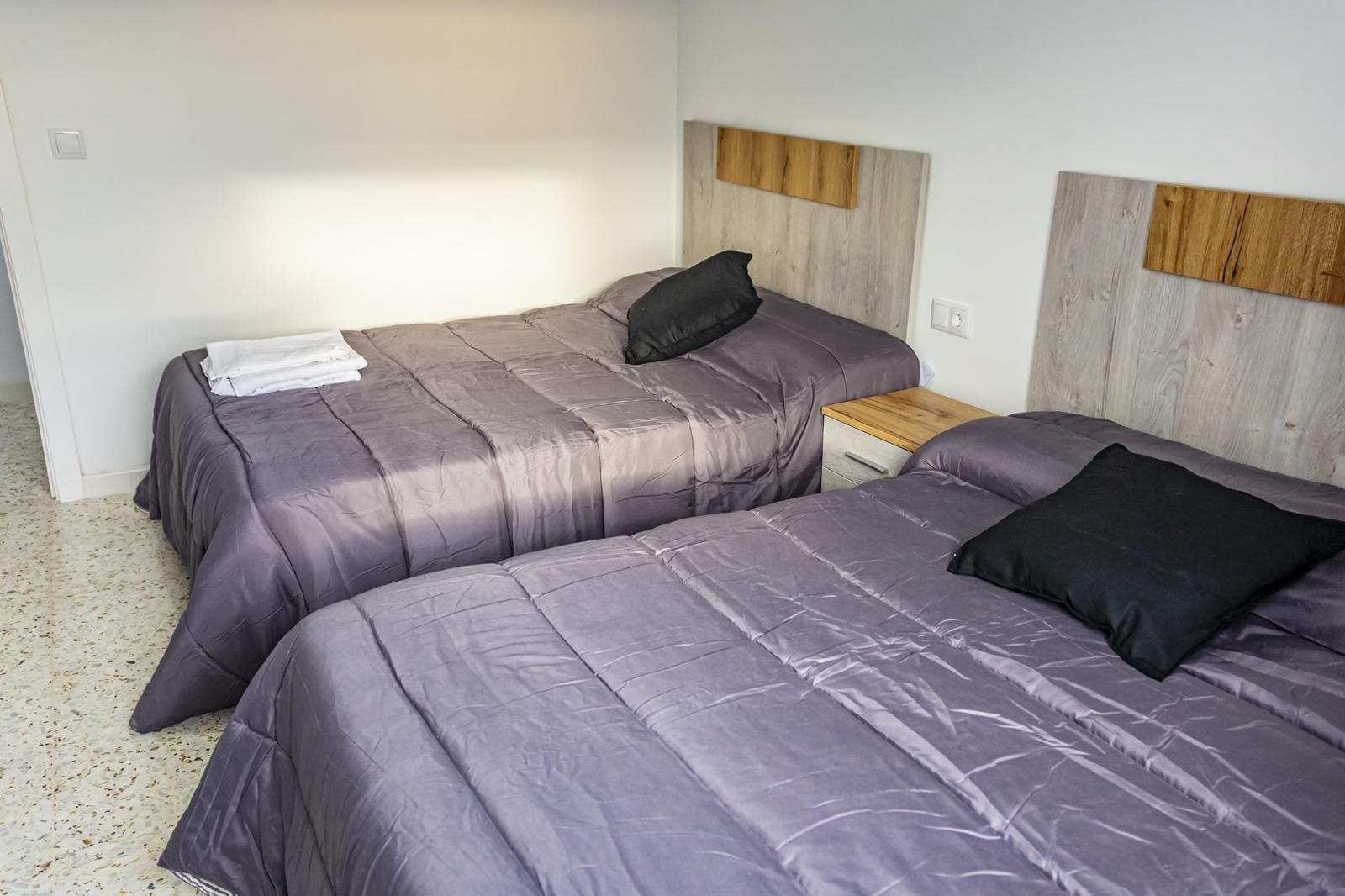 Dos camas individuales en una habitación con edredones grises, almohadas negras y cabeceros de madera.