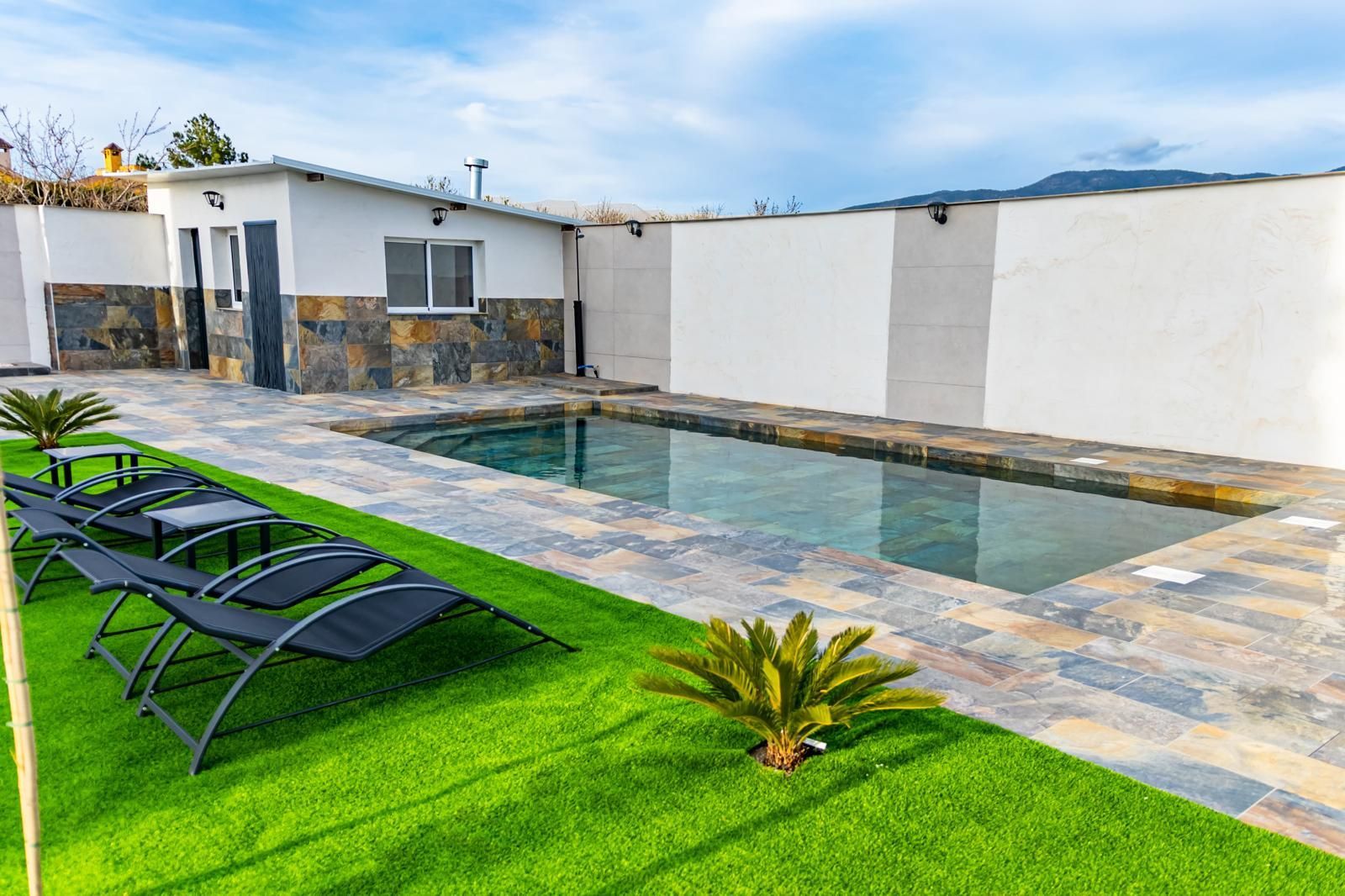 Zona de piscina con tumbonas, piscina pequeña y paredes blancas. Césped verde alrededor de la piscina.