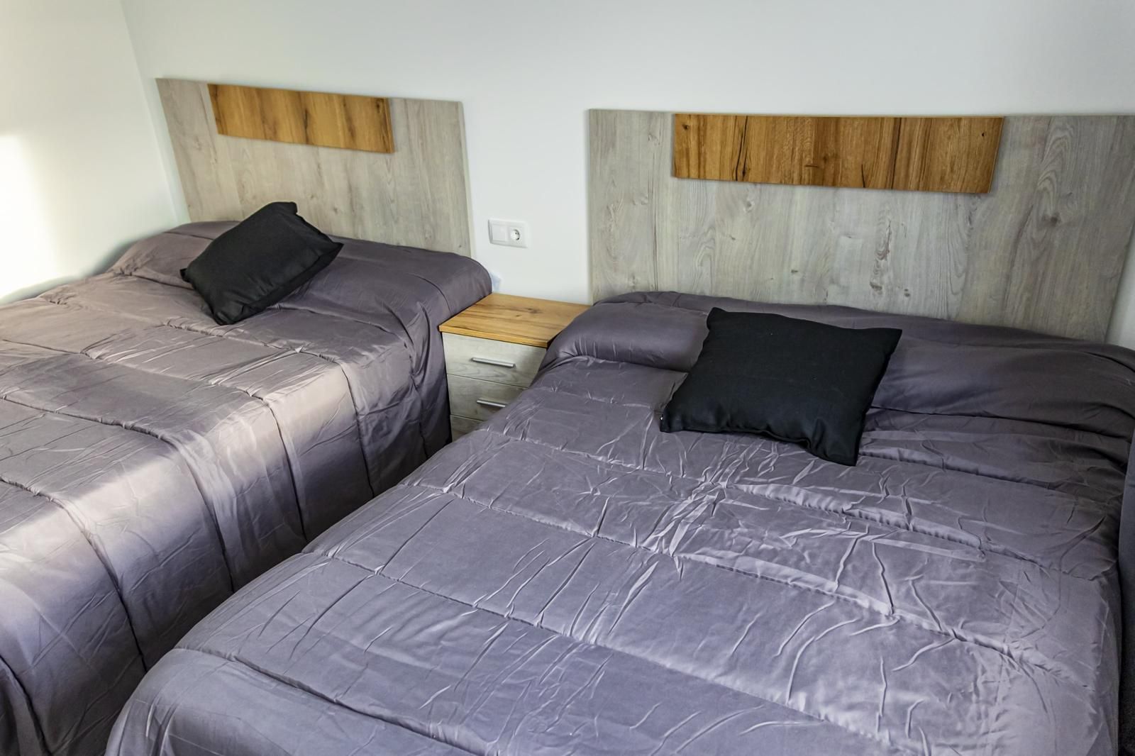 Dos camas con ropa de cama gris, almohadas negras y cabeceros de madera en una habitación.