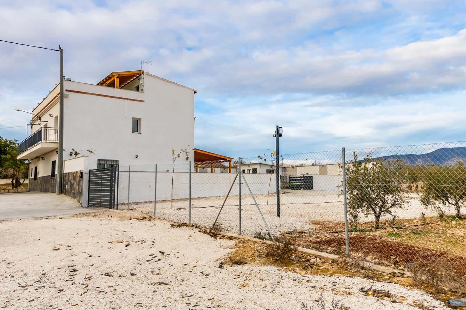 Edificio blanco con valla y terreno árido bajo un cielo nublado.