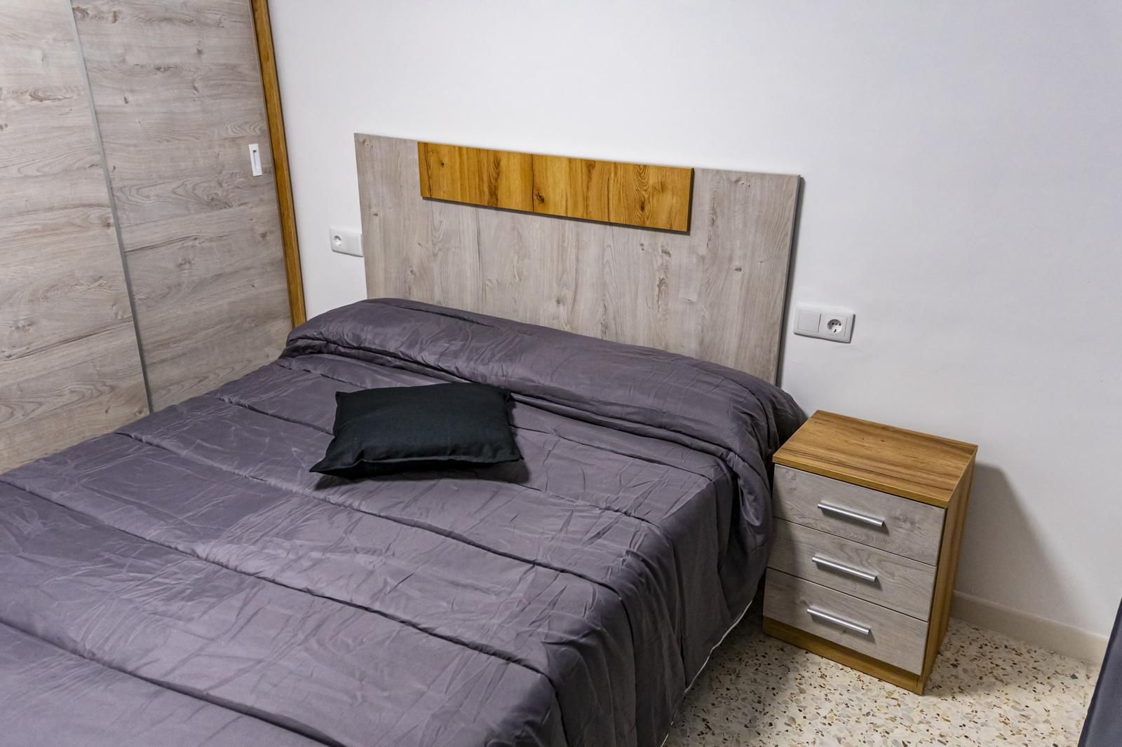 Dormitorio con cama, mesita de noche y armario. La cama tiene ropa de cama gris, almohada negra y cabecero de madera.