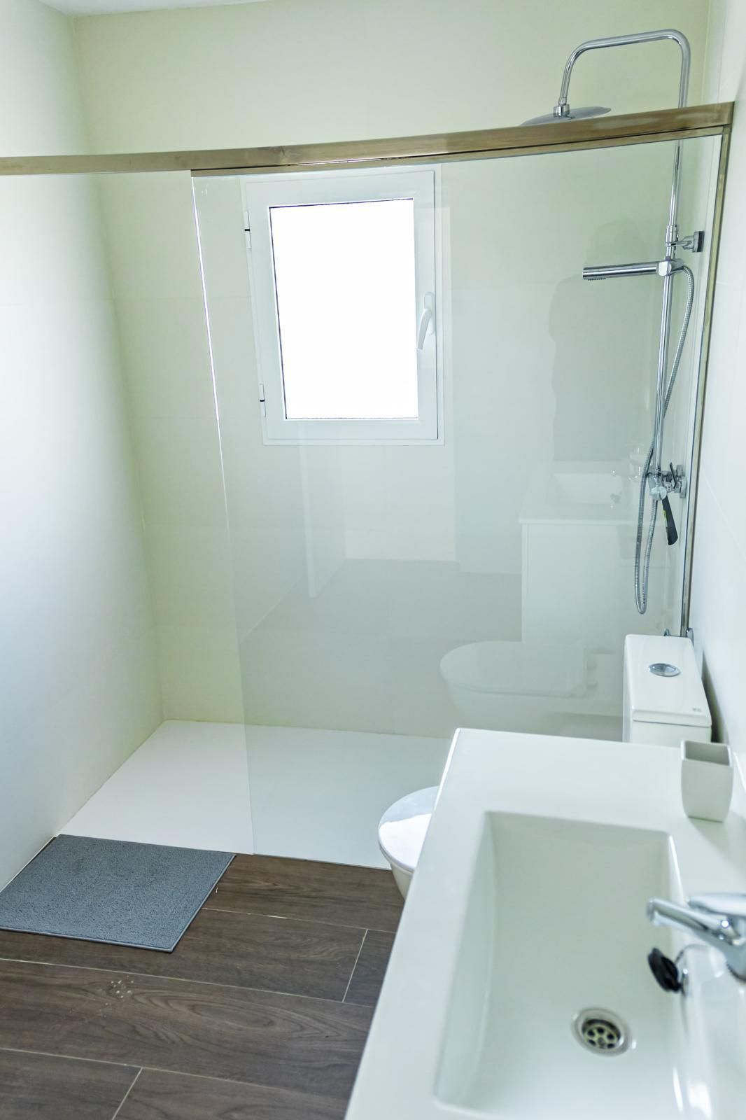 Pequeño baño blanco con ducha de cristal, inodoro y lavabo; suelo de madera oscura y ventana.