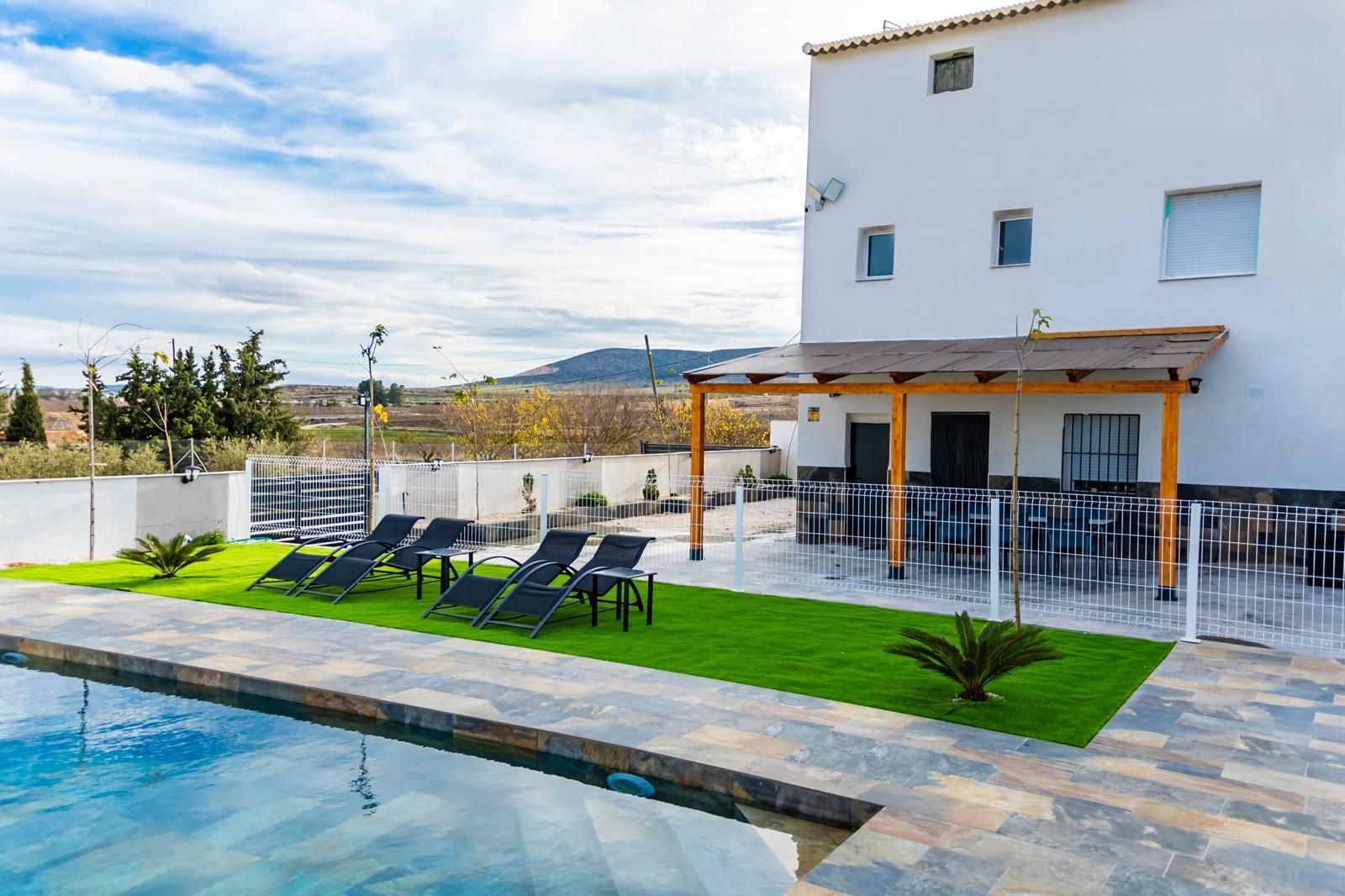 Vista a la piscina: edificio blanco, piscina, tumbonas, patio con mesas, soleado y cielo azul.