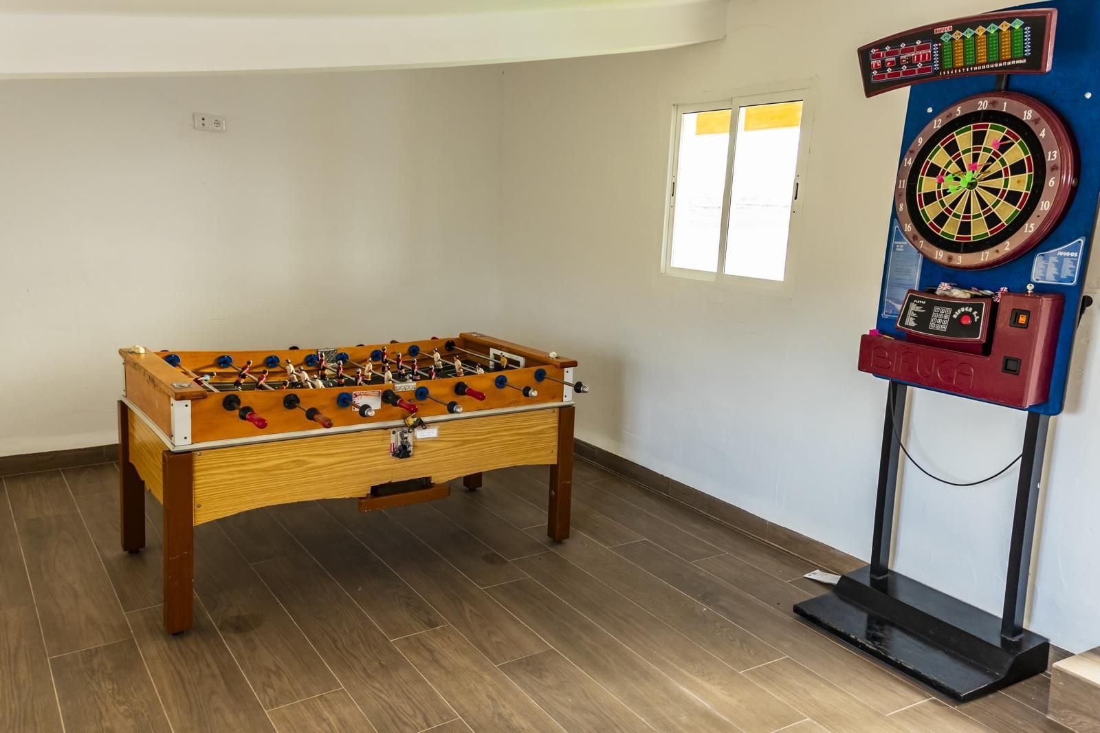 Mesa de futbolín y diana electrónica en una sala con suelos de madera clara y paredes blancas.