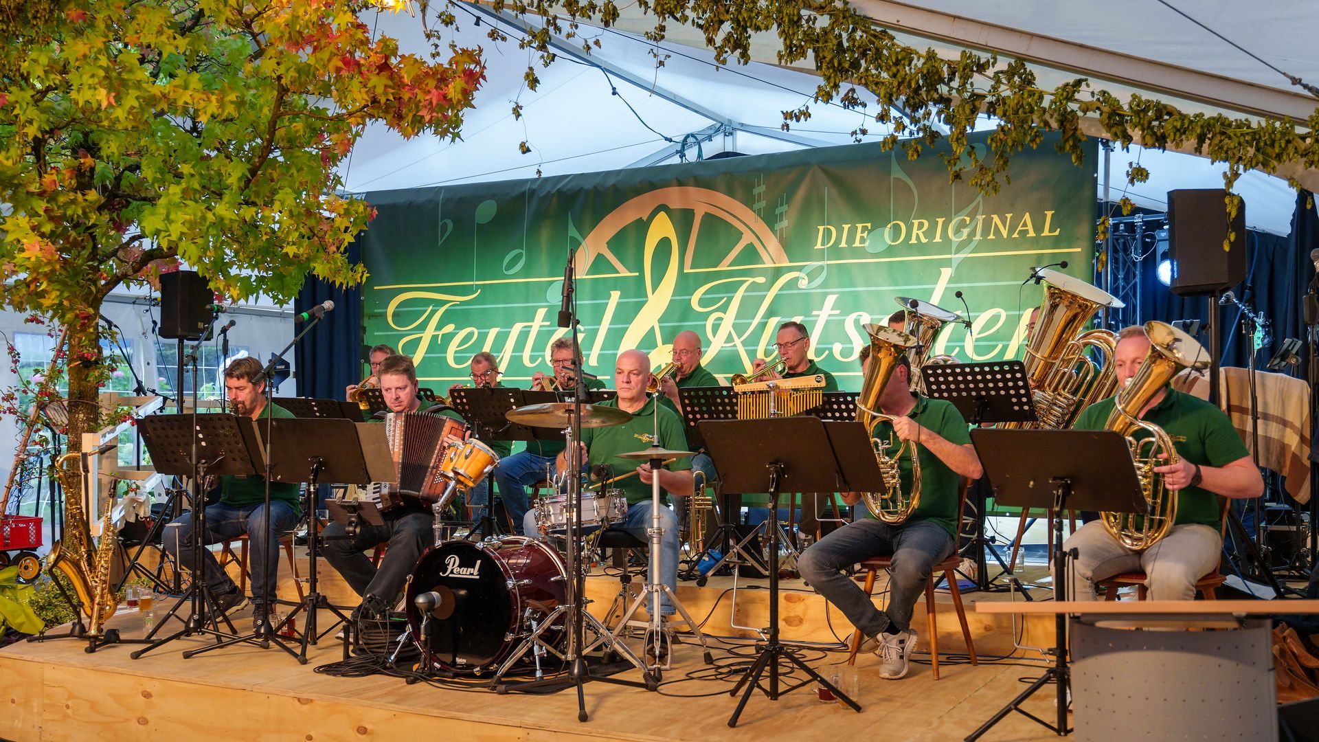 Eine Jazzband spielt im Freien auf einer Bühne unter Bäumen in der Abenddämmerung; grüner Hintergrund, Instrumente gut sichtbar.