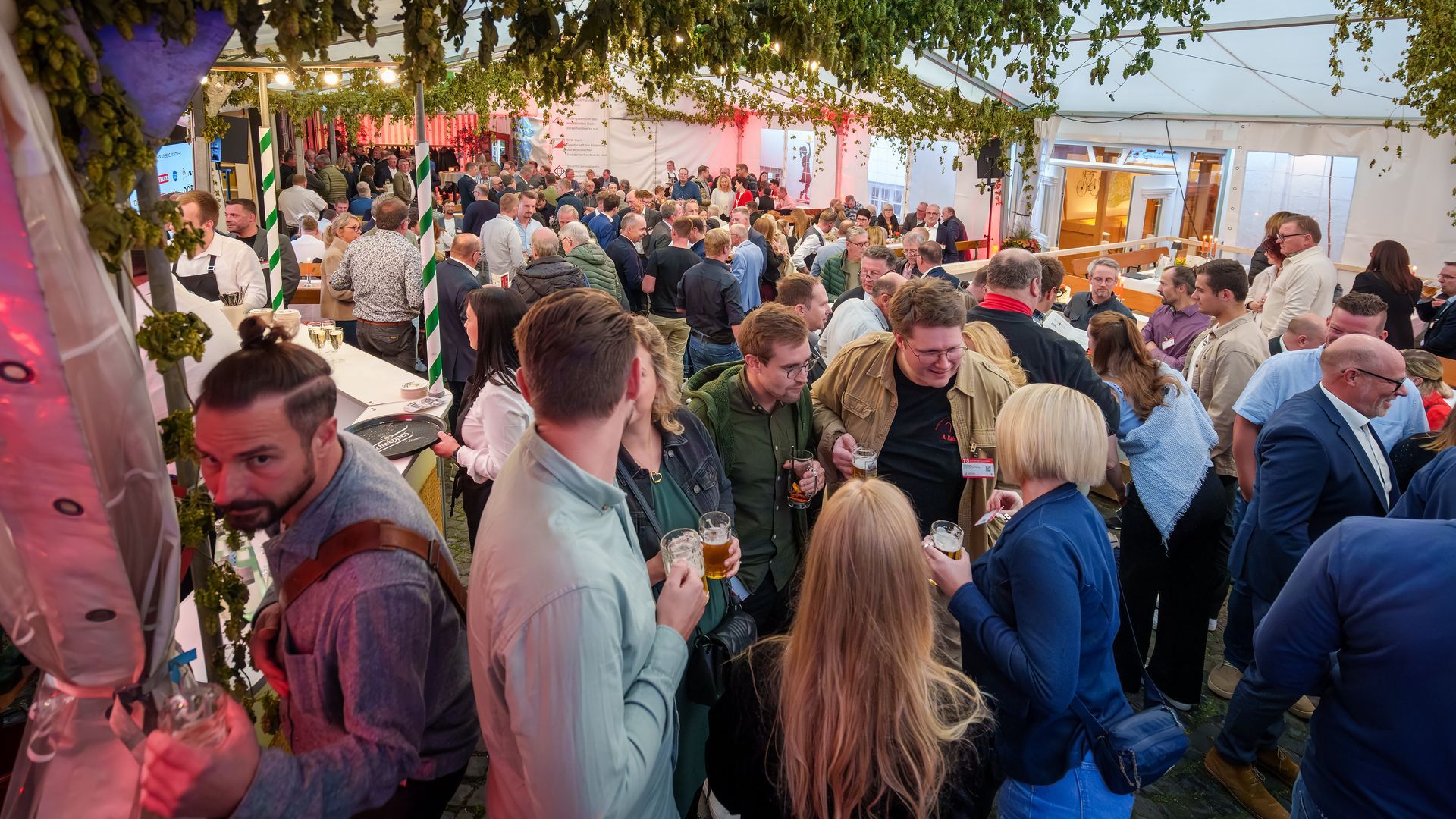 Eine gut besuchte abendliche Open-Air-Party unter Lichterketten, die Leute mischen sich bei Getränken an Tischen und in einem Innenhof.