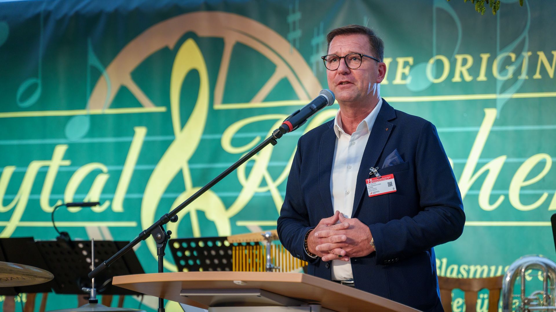 Mann spricht an einem Rednerpult auf einer Bühne vor grünem und goldenem Hintergrund und Veranstaltungsschildern.