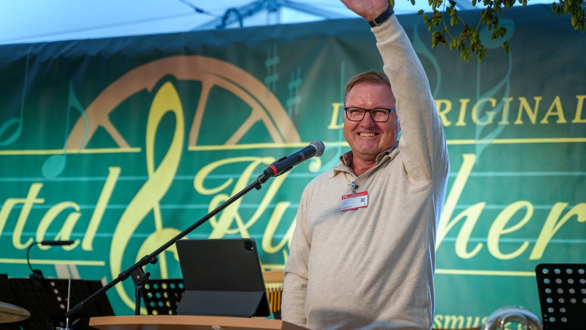 Ein Mann spricht auf der Bühne, hebt einen Arm; vor einem blauen Banner stehen ein Laptop und ein Mikrofon.