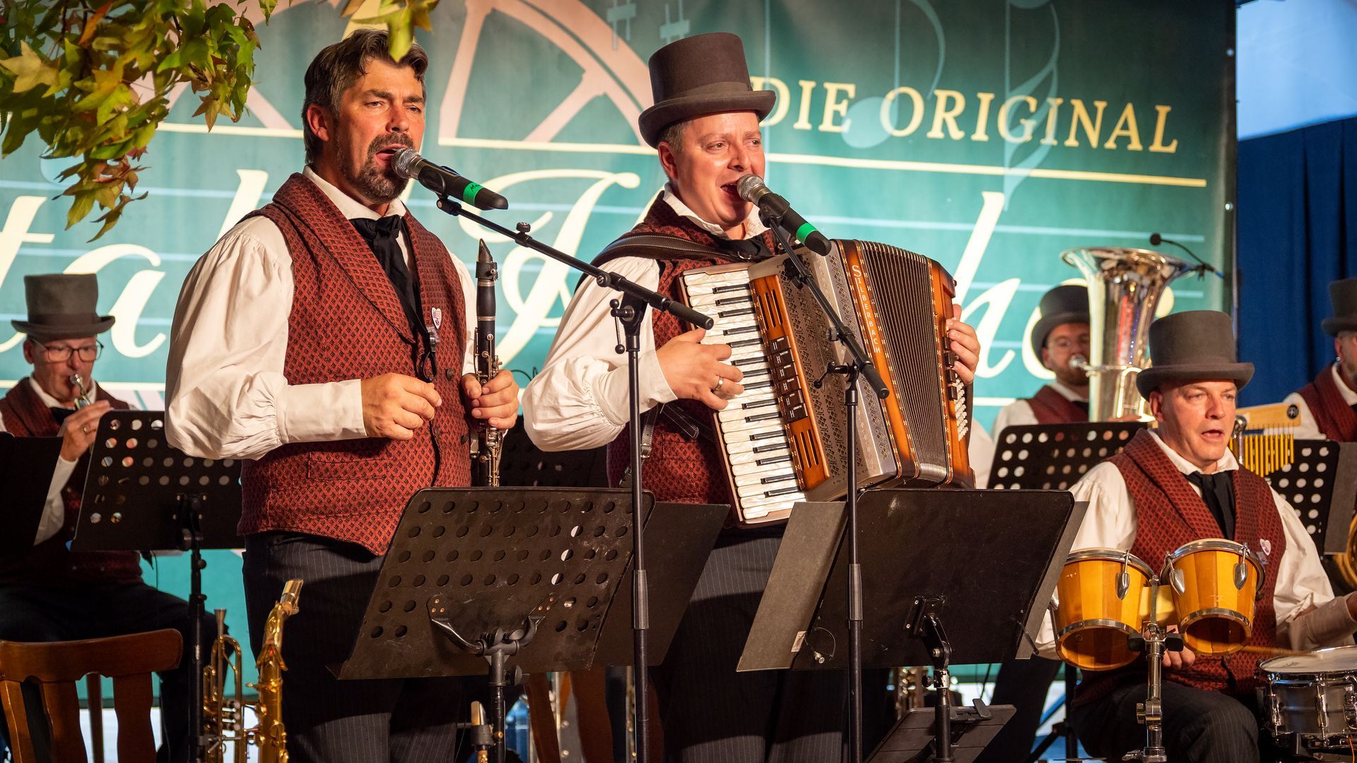 Musiker spielen auf der Bühne mit Akkordeon, Gitarre und Banjo vor einem türkisfarbenen Hintergrund.