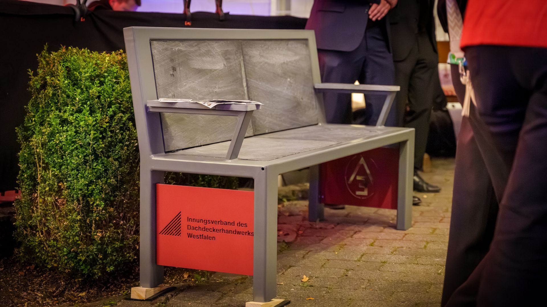 Graue Metallbank mit roten Seitenwänden, die nachts draußen auf einer Rasenfläche steht, in der Nähe von Menschen.