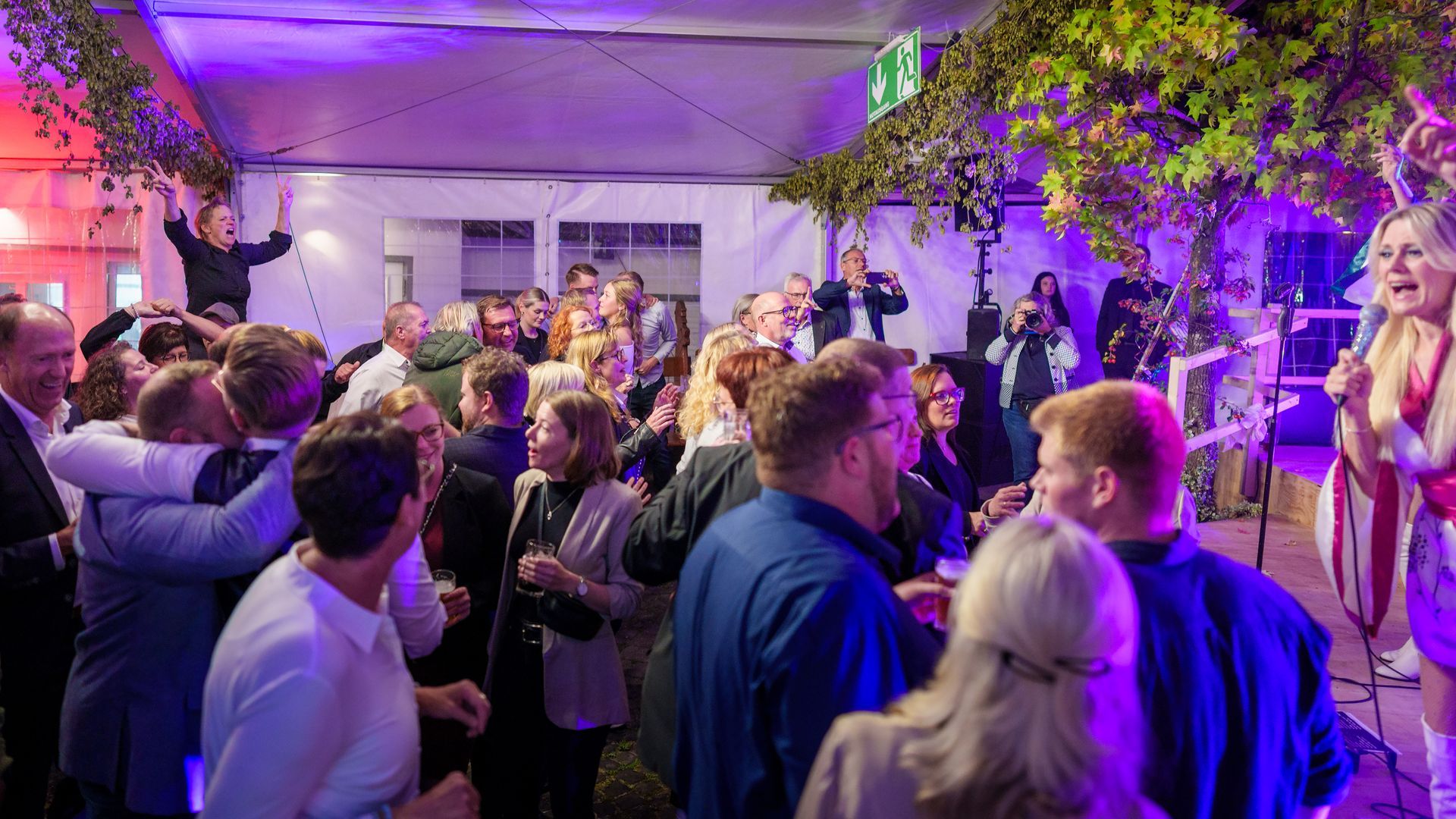 Eine überfüllte Indoor-Party mit tanzenden Gästen, violetter Beleuchtung und einem Sänger auf einer kleinen Bühne.