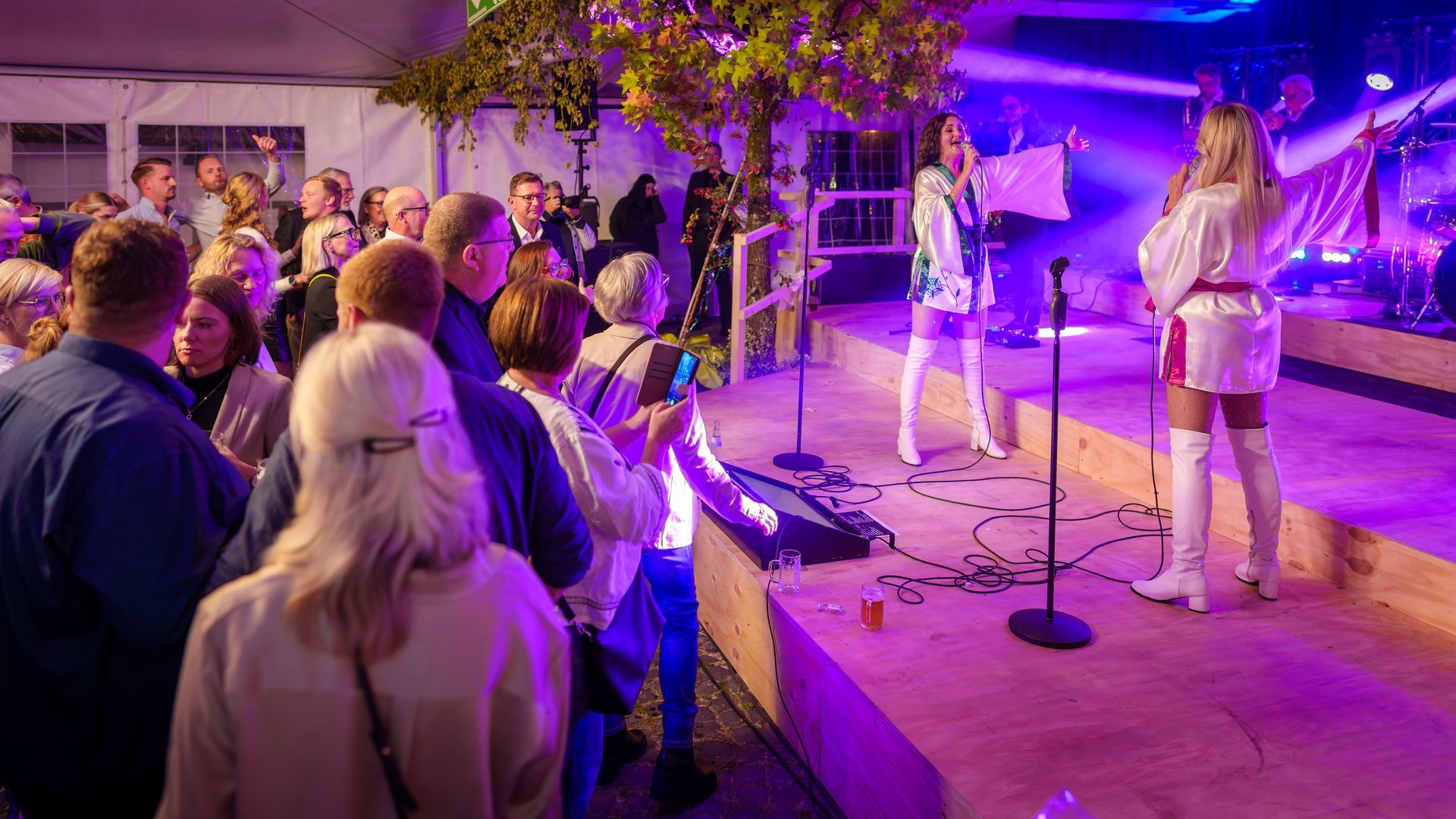 Eine Live-Band spielt auf einer violett beleuchteten Bühne bei einer Indoor-Veranstaltung, das Publikum schaut in der Nähe zu.