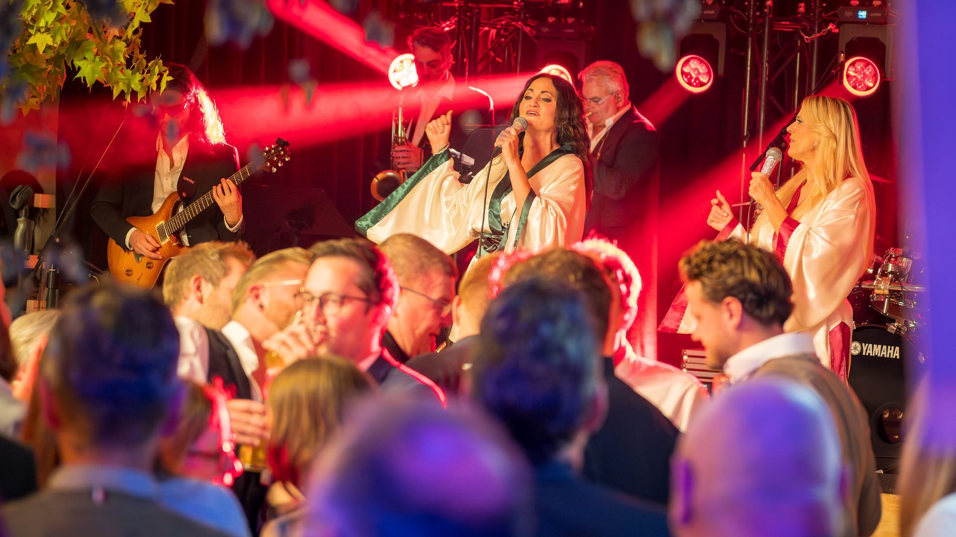 Eine Live-Band spielt auf einer warm beleuchteten Bühne vor einem gut besuchten Publikum bei einer Indoor-Veranstaltung.
