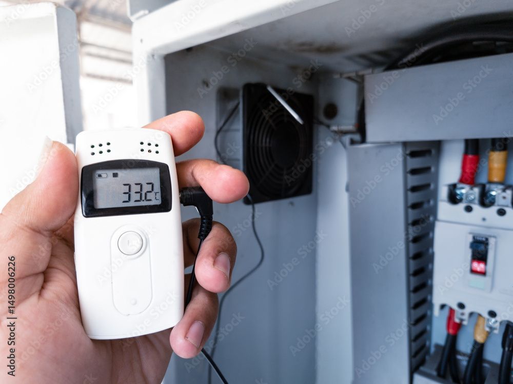 Hand hält ein digitales Thermometer mit der Anzeige 33,2 in einem Schaltschrank mit Kabeln und einem Ventilator.