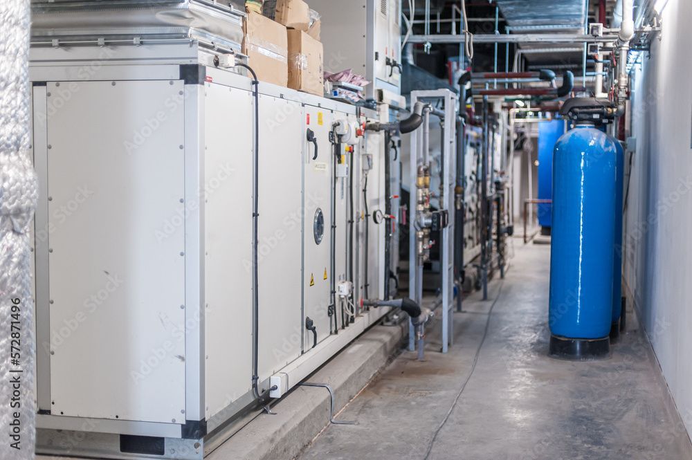 Ein schmaler Versorgungskorridor mit großen Industrieanlagen und blauen Tanks. Die Maschinen sind überwiegend weiß und grau, Rohre und Kabel sind sichtbar.