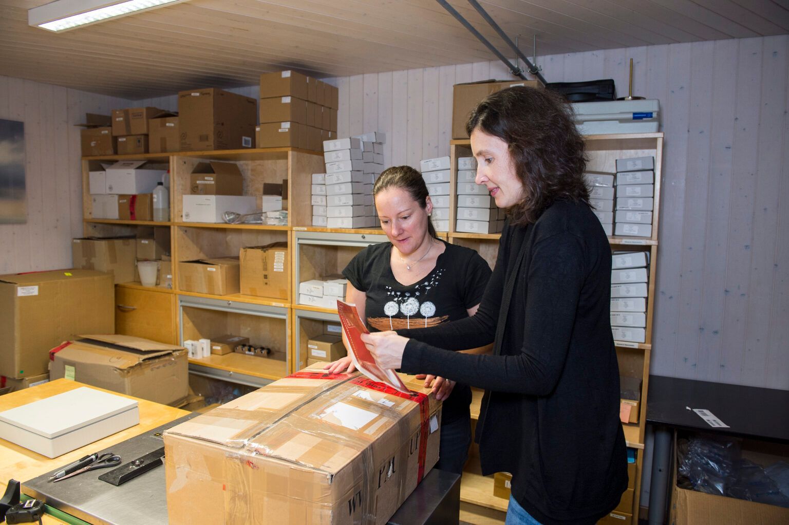 Zwei Frauen verpacken in einem Lagerraum einen großen Karton. Eine lächelt, die andere liest eine Karte. Im Hintergrund stehen Regale voller Kartons.