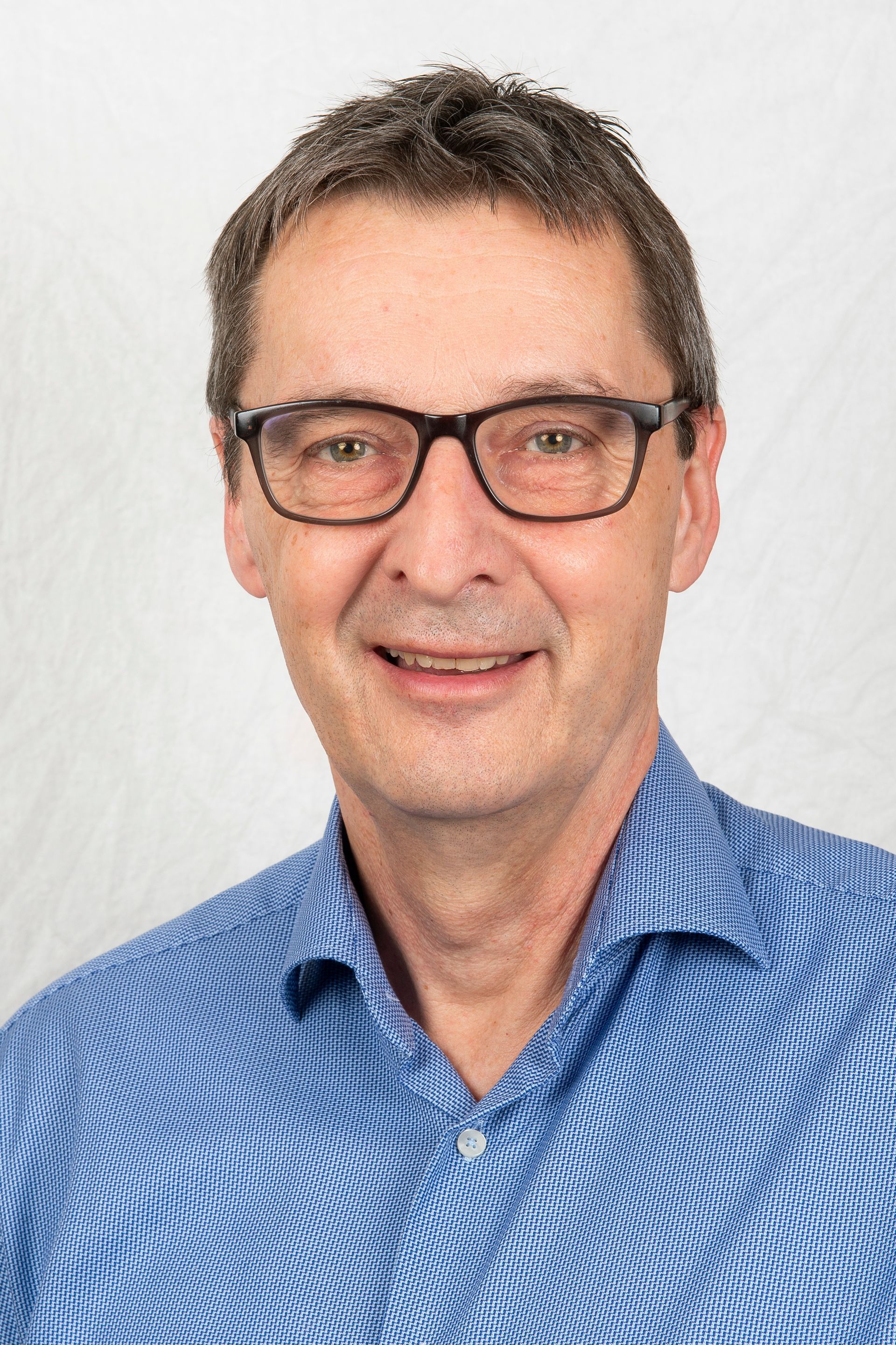 Mann mit kurzen braunen Haaren und blau gemustertem Hemd lächelt vor neutralem Hintergrund in die Kamera.