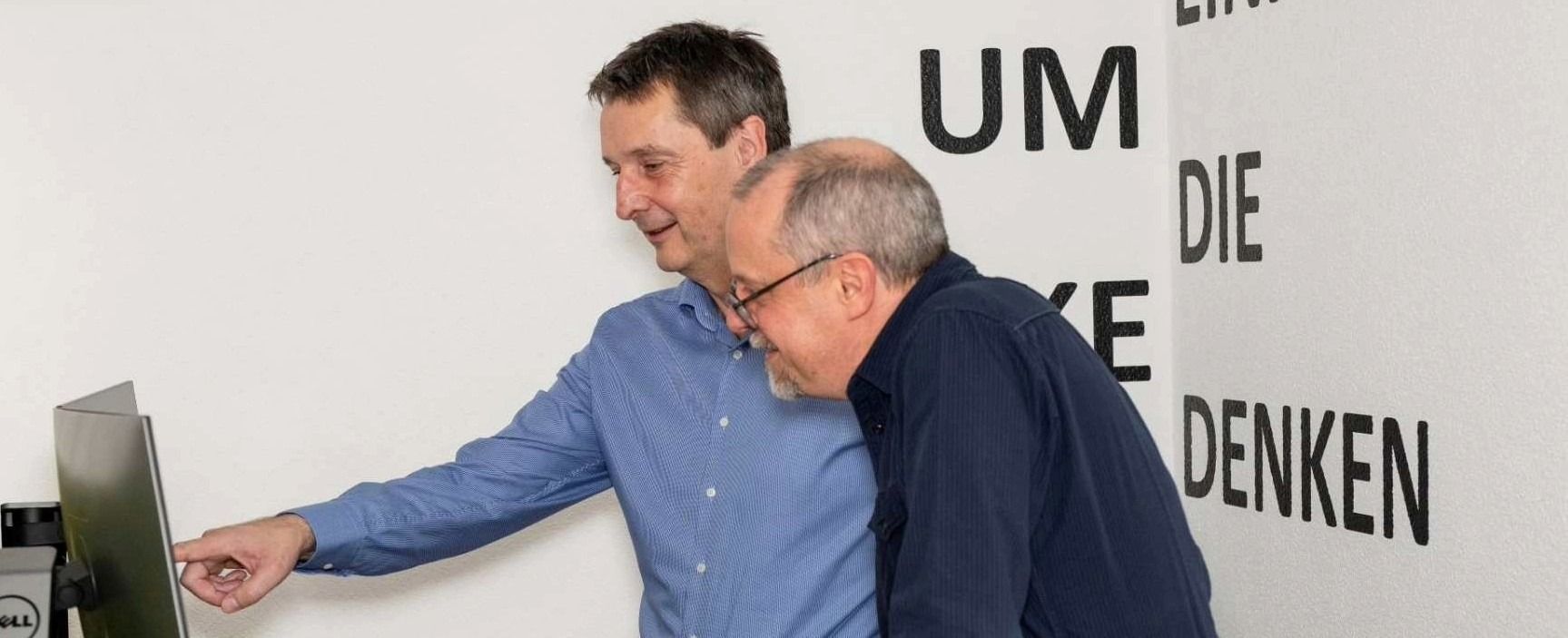 Zwei Männer schauen auf einen Computerbildschirm. Einer zeigt auf den Bildschirm, der andere trägt eine Brille und lächelt. Weiße Wand mit deutschem Text im Hintergrund.