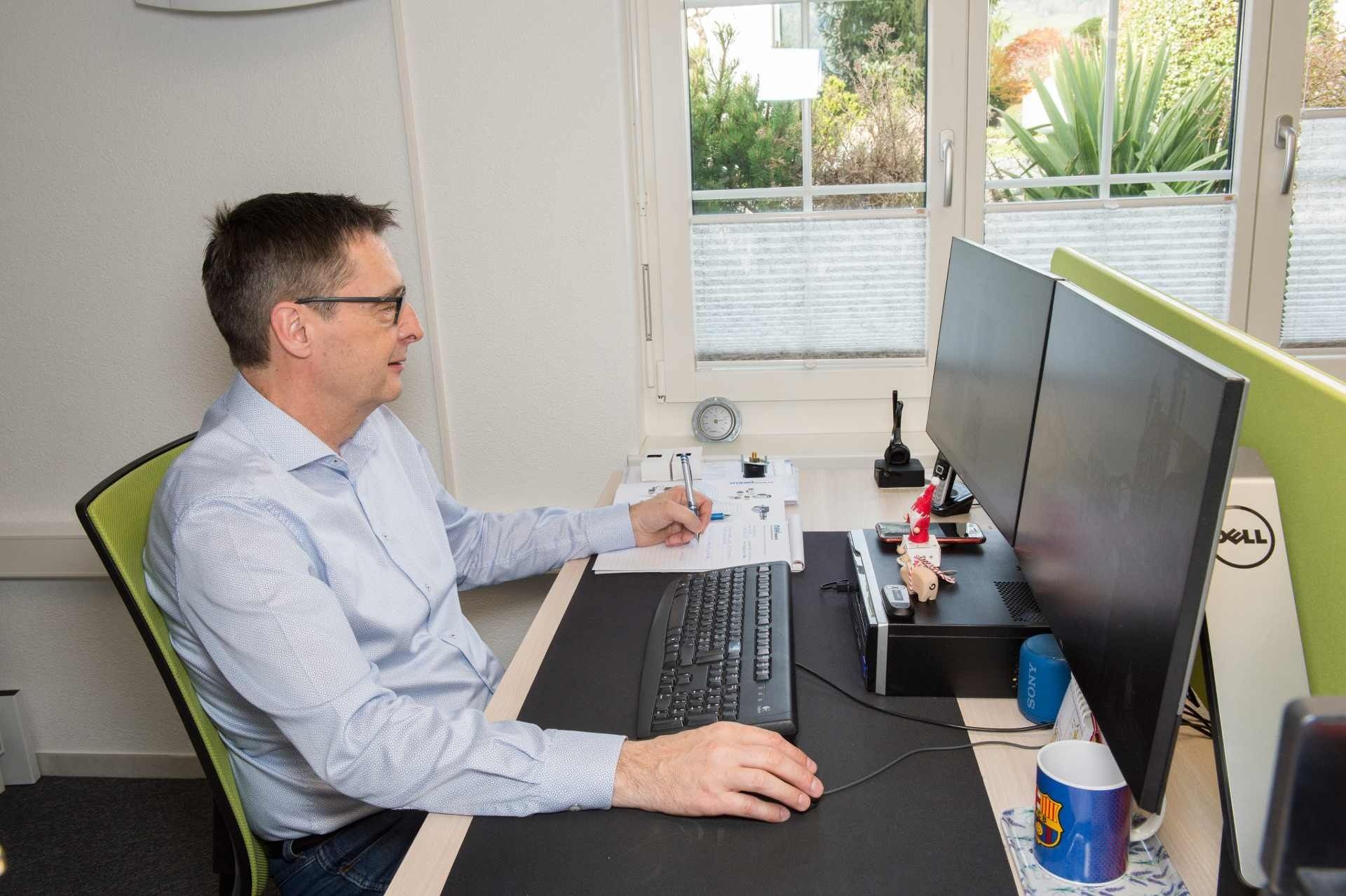 Mann sitzt an einem Schreibtisch mit zwei Monitoren und tippt und schreibt. Er trägt eine Brille und ein hellblaues Hemd und arbeitet in einem gut beleuchteten Büro.