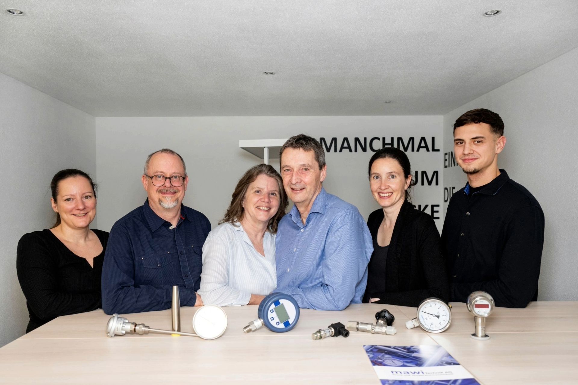 Eine Gruppe von sechs lächelnden Personen hat sich hinter einem Tisch mit Industriemessgeräten versammelt. Sie befinden sich in einem hell erleuchteten Büroraum.