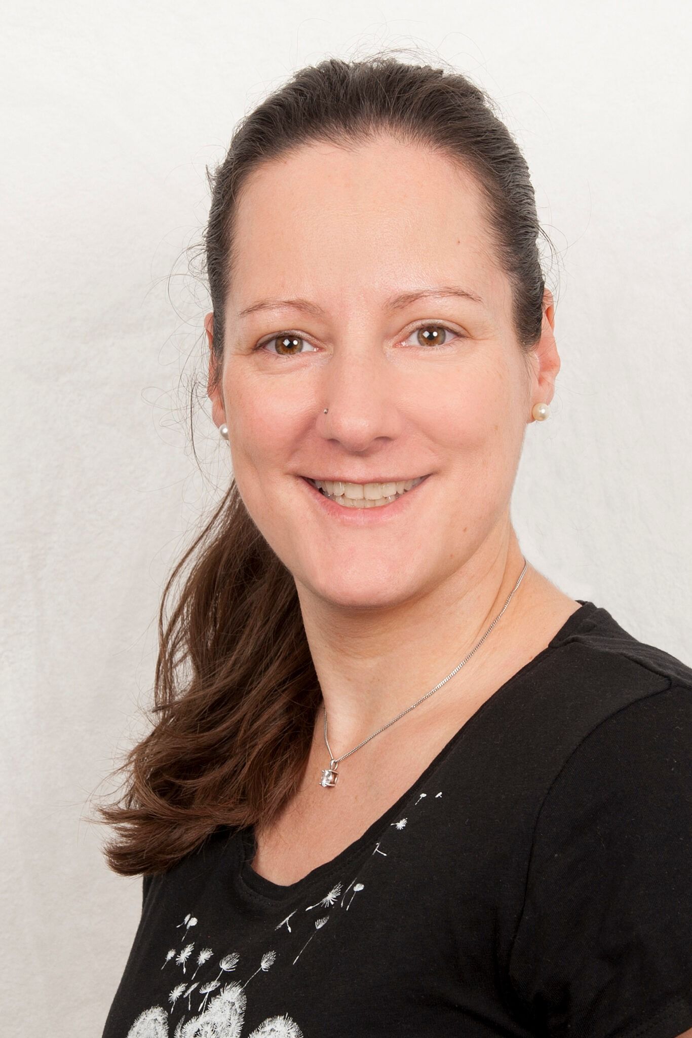 Frau mit braunem Haar zu einem Pferdeschwanz, lächelnd, mit schwarzem Hemd und Halskette vor weißem Hintergrund.