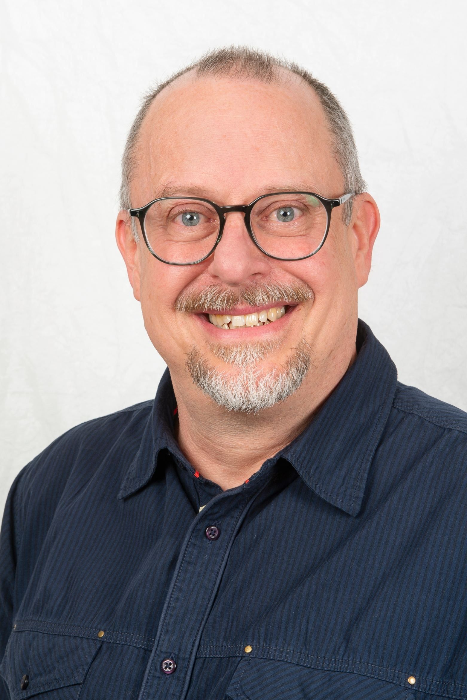 Mann mit Brille und ergrauendem Bart lächelt, trägt ein blaues Button-Down-Hemd vor einem weißen Hintergrund.