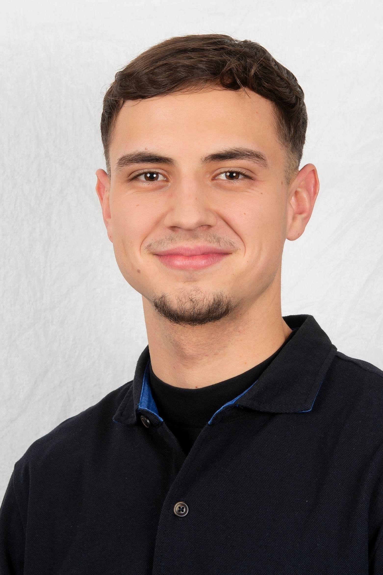 Lächelnder junger Mann mit kurzen braunen Haaren und Spitzbart, bekleidet mit einem dunkelblauen Hemd und einem schwarzen Unterhemd vor einem schlichten weißen Hintergrund.