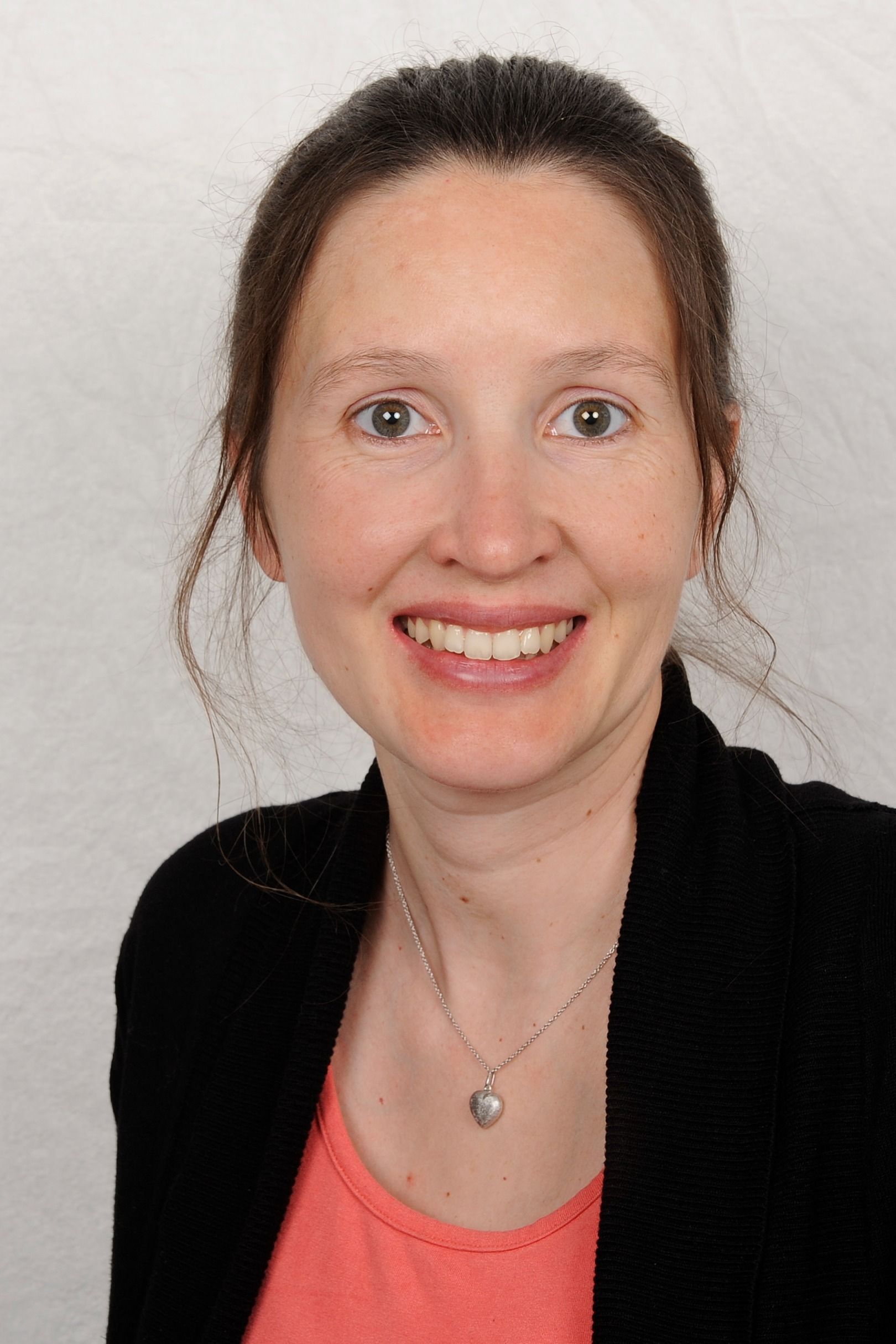 Eine Frau mit heller Haut lächelt und trägt eine schwarze Jacke über einem rosa Hemd. Sie hat zurückgekämmtes braunes Haar und eine silberne Halskette.