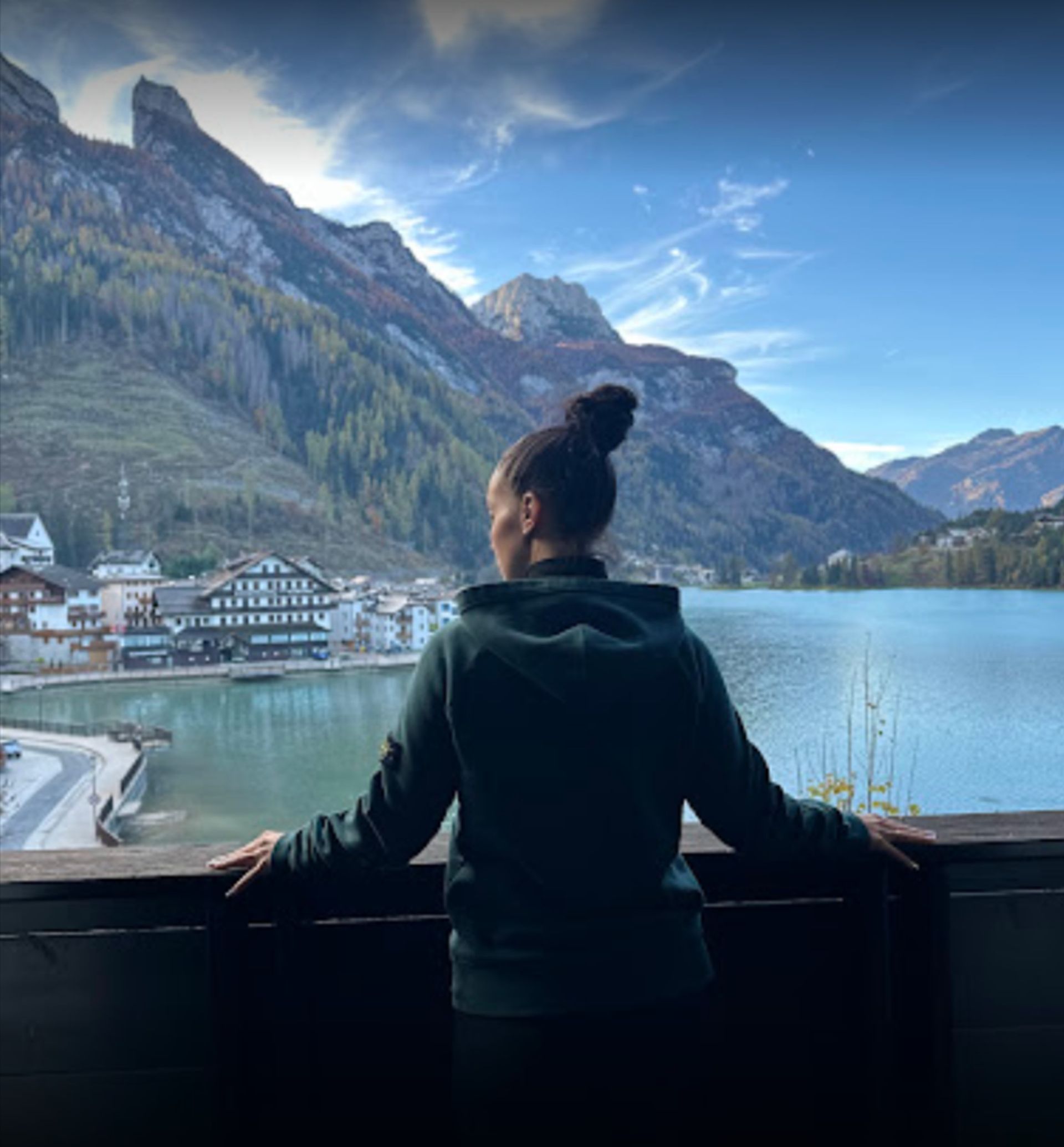 Persona che guarda un lago e le montagne, con le braccia appoggiate su una ringhiera di legno. Sullo sfondo un villaggio.