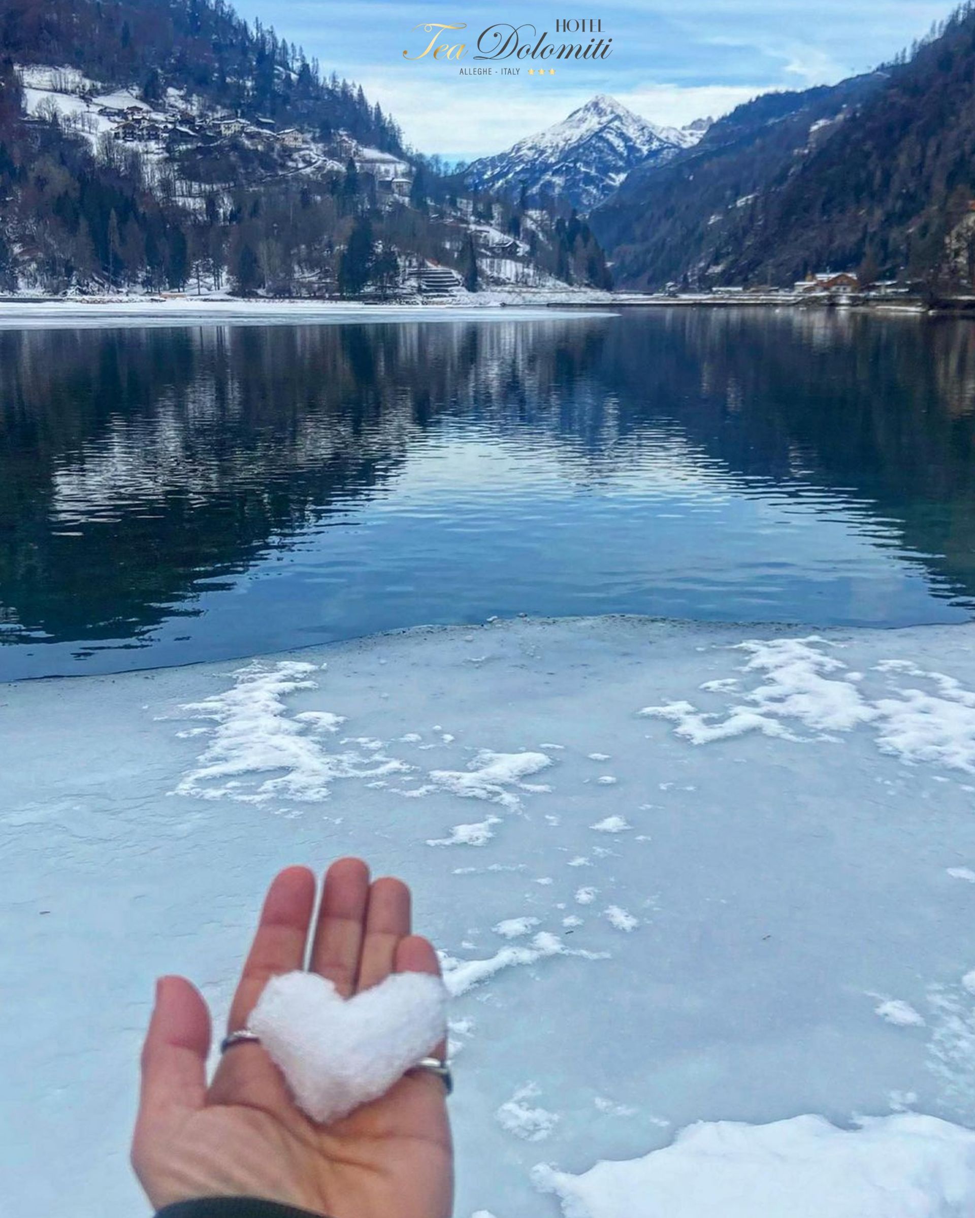 Mano che tiene un cuore di neve, con un lago innevato e montagne sullo sfondo.
