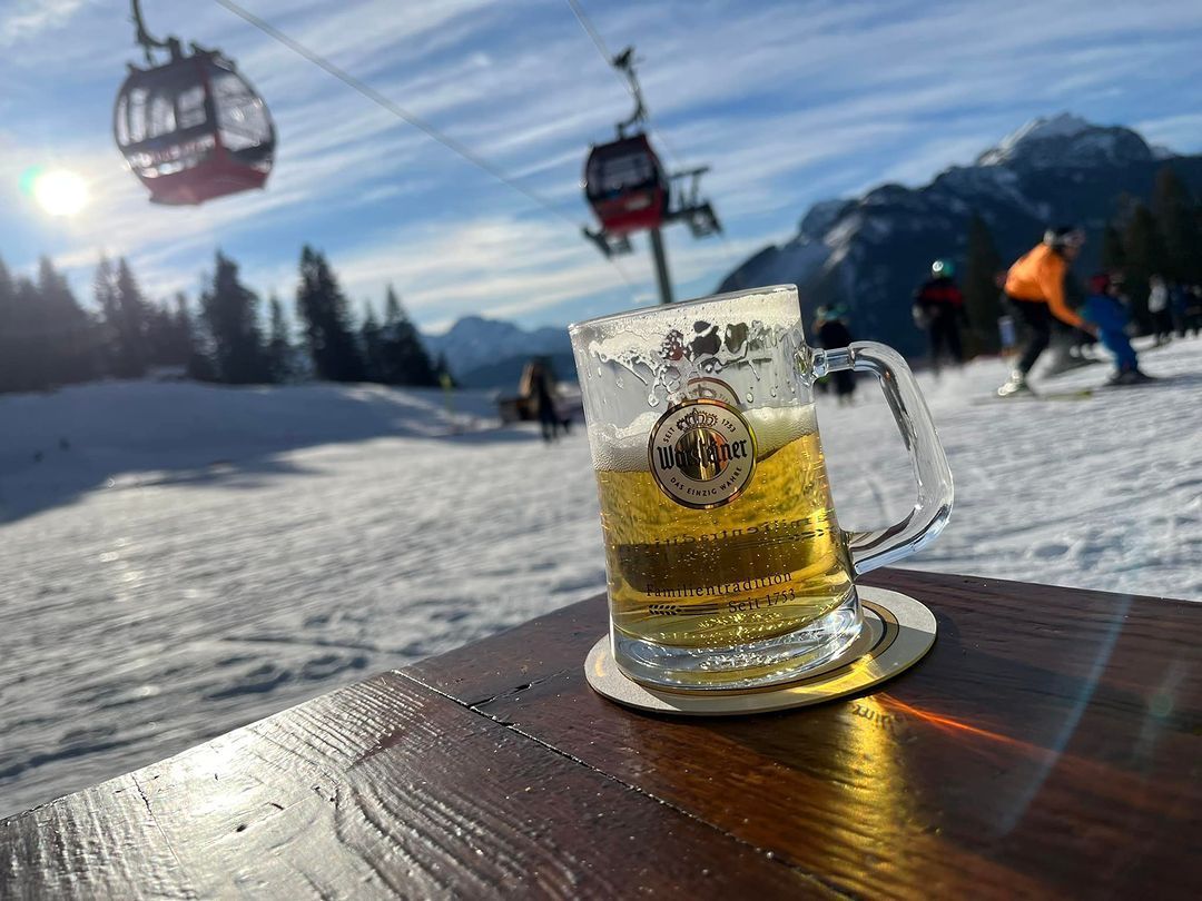Boccale di birra sul tavolo in una stazione sciistica; sciatori, funivia, montagne innevate sullo sfondo. Giornata di sole.