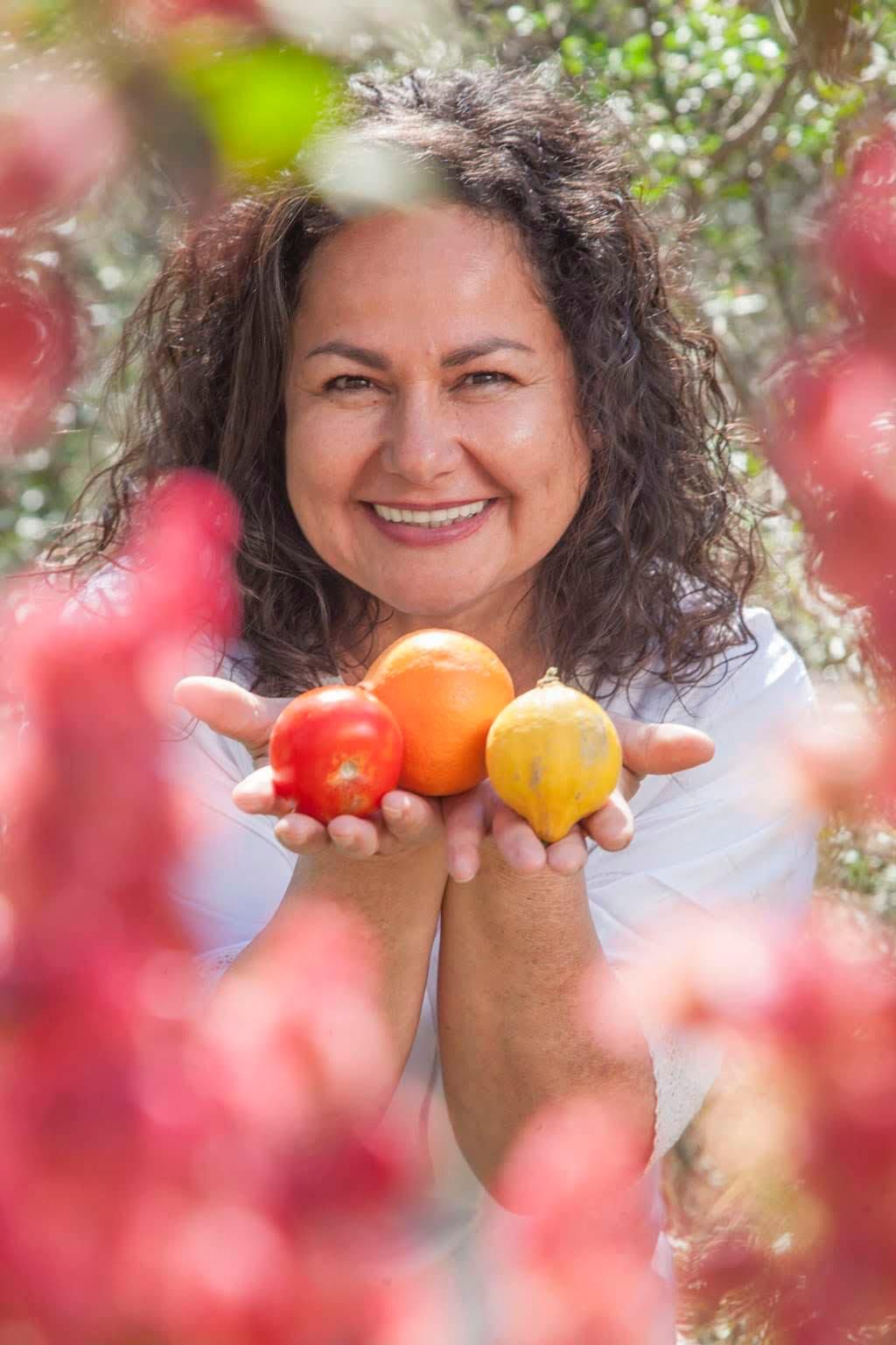Mujer sonriendo, sosteniendo tomate, naranja y limón al aire libre, rodeada de flores rosas.